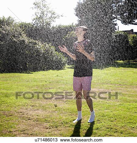 Stock Image - Young man standing in garden sprinkler. Fotosearch