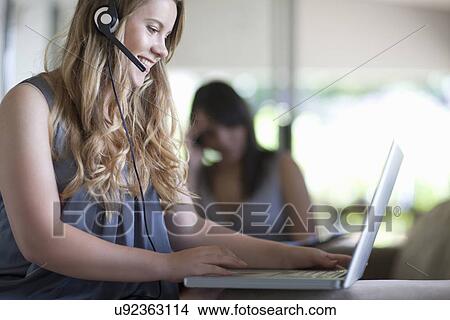 Picture - Young office worker talking on headset whilst on laptop. Fotosearch