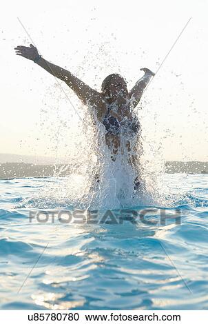 Stock Image - Young woman with arms out splashing in swimming pool. Fotosearch