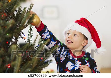 Stock Photo - Boy (6-7) with santa hat decorating Christmas tree. Fotosearch