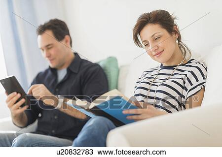 Stock Image - Couple sitting on sofa, woman reading book. Fotosearch