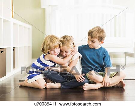 Stock Image - Girls (4-5, 6-7) and boy (8-9) sitting on floor and embracing. Fotosearch