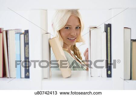 Stock Image - Portrait of young woman peeking through bookshelf. Fotosearch
