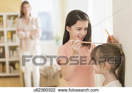 Stock Image - Sisters (4-5, 8-9) measuring height, mother watching. Fotosearch