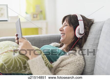 Stock Image - Woman relaxing on sofa, listening to music on tablet pc. Fotosearch