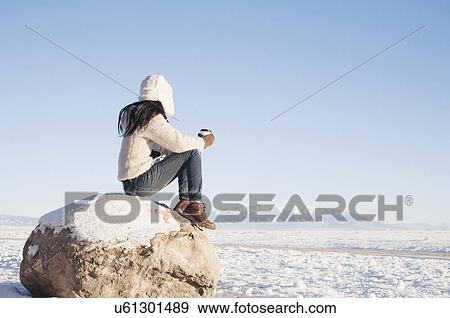 Stock Photo - Woman sitting on rock with cup in winter. Fotosearch