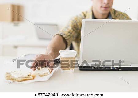 Stock Image - Young man eating sandwich and using laptop. Fotosearch