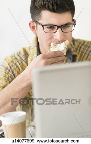 Stock Photo - Young man eating sandwich and using laptop. Fotosearch