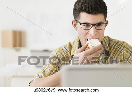 Stock Photo - Young man eating sandwich and using laptop. Fotosearch