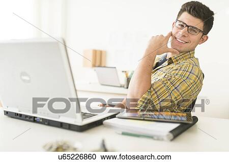 Stock Image - Young man sitting in front of desk and smiling. Fotosearch