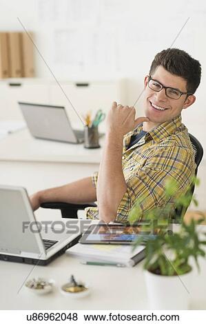 Stock Photo - Young man sitting in front of desk and smiling. Fotosearch