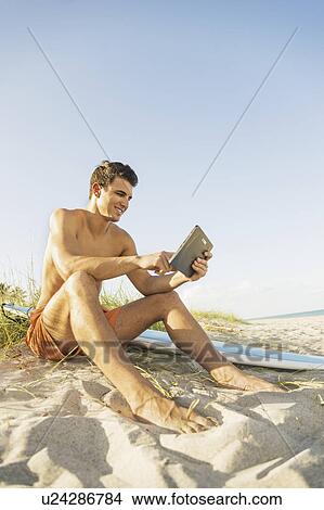 Young man sitting on beach with surfboard and digital tablet View Large Photo Image Picture - Young man sitting on beach with surfboard and digital tablet. Fotosearch