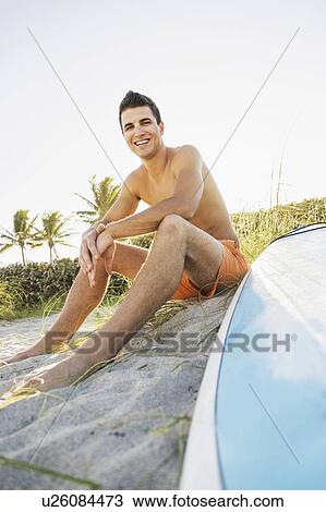Stock Image - Young man sitting on beach with surfboard. Fotosearch