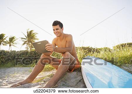 Young man sitting on beach with surfboard and digital tablet View Large Photo Image Stock Photo - Young man sitting on beach with surfboard and digital tablet. Fotosearch
