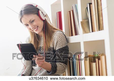 Stock Image - Young woman sitting on floor and listening to music on digital tablet. Fotosearch