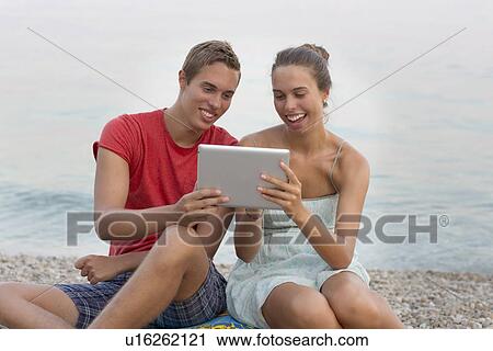 Stock Image - Brother and sister sitting on beach, using digital tablet. Fotosearch