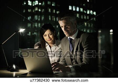 Stock Image - Business colleagues working late in office on laptop. Fotosearch