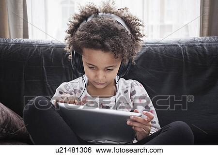Stock Photograph - Girl sitting on sofa using digital tablet and headphones. Fotosearch
