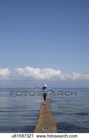 Stock Image - Girl walking along narrow pier at coast. Fotosearch