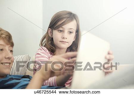 Picture - Sister and older brother on sofa using digital tablet. Fotosearch