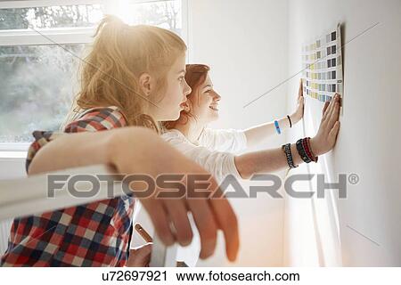 Stock Image - Teenage sisters holding color swatch against wall. Fotosearch