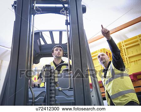Stock Image - Trainer instructs male apprentice on forklift truck. Fotosearch