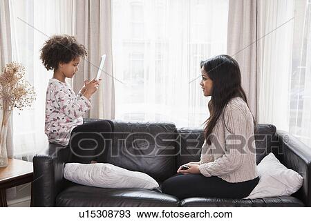 Stock Image - Young girl taking photograph of teenage sister on digital tablet. Fotosearch
