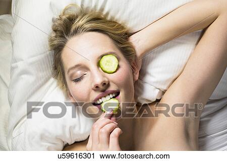 Stock Image - Young woman having facial, biting slice of cucumber. Fotosearch