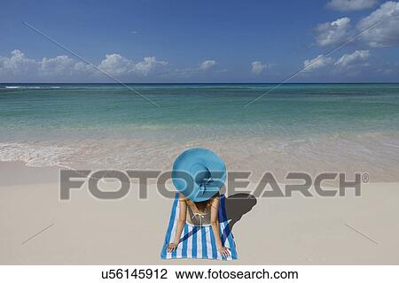 Stock Image - Young woman relaxing on beach with blue sunhat. Fotosearch