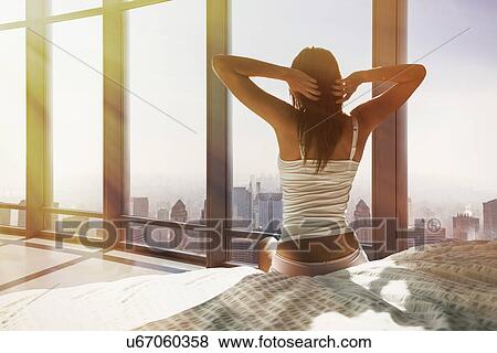 Stock Photo - Young woman sitting on bed, stretching, overlooking city. Fotosearch