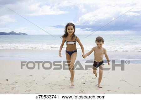 Stock Image - Brother and sister running on beach, holding hands. Fotosearch