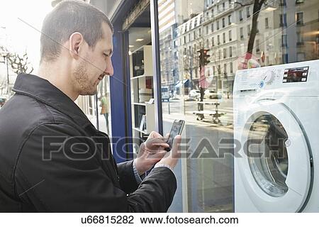 Stock Image - Mid adult man checking out washing machine in shop using smartphone. Fotosearch