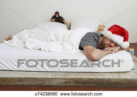 Stock Image - Mid adult man in santa hat asleep in bed. Fotosearch