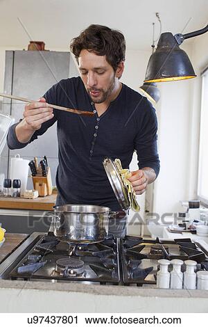 Stock Image - Mid adult man tasting food from wooden spoon in kitchen. Fotosearch