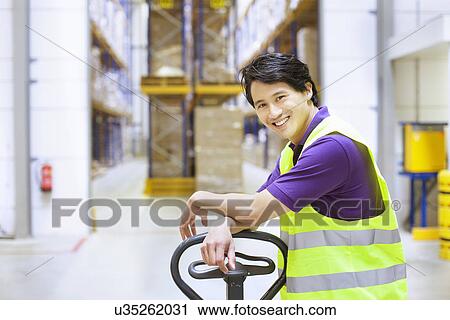 Stock Image - Portrait of male worker leaning on pallet jack in distribution warehouse. Fotosearch