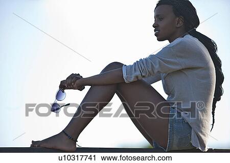 Stock Image - Portrait of young woman sitting, holding sunglasses. Fotosearch