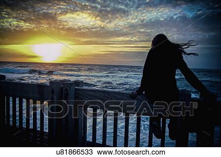 Stock Image - Silhouette of young woman sitting on sea front fence at dawn. Fotosearch