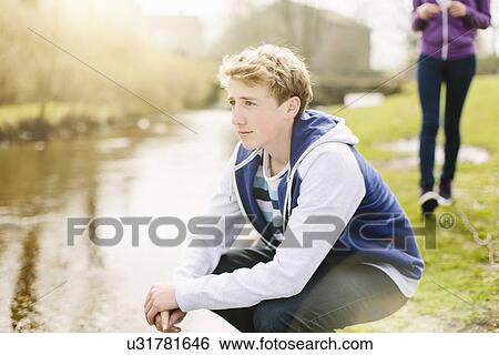 Stock Photograph - Unhappy teenage boy crouching on riverbank. Fotosearch