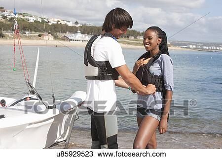 Stock Image - Young adult friends checking life jackets on waterfront. Fotosearch