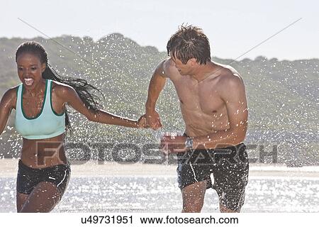 Stock Image - Young couple holding hands running through sea. Fotosearch