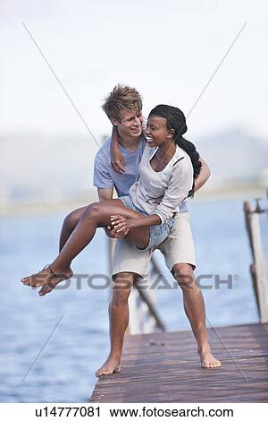 Stock Image - Young couple of jetty, man lifting woman. Fotosearch