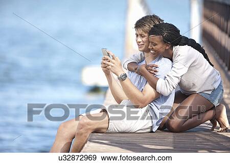 Stock Photo - Young couple sitting on jetty, looking at mobile phone. Fotosearch