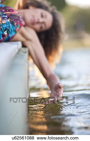 Stock Photo - Young woman laying on jetty, hand touching water. Fotosearch