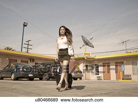 Stock Photo - Young woman pulling wheeled suitcase outside motel. Fotosearch