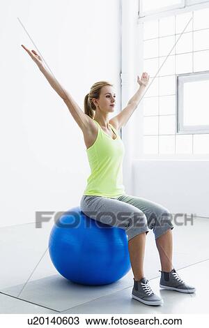 Stock Image - Young woman sitting on exercise ball, arms raised. Fotosearch