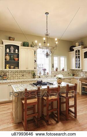 Stock Photograph - Lit chandelier hanging over high stools at kitchen island at home; Santa Fe; New Mexico; USA. Fotosearch