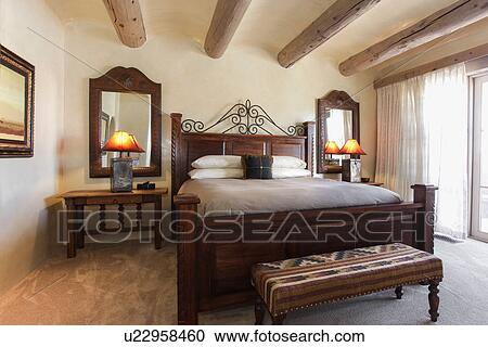 Stock Image - Lit table lamps by tidy bed with wooden beam ceiling in bedroom at home; Santa Fe; New Mexico; USA. Fotosearch