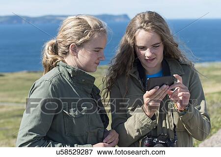 Stock Photo - Girls looking at mobile phone on coast, Cape Spear, St. John's, Newfoundland And Labrador, Canada. Fotosearch