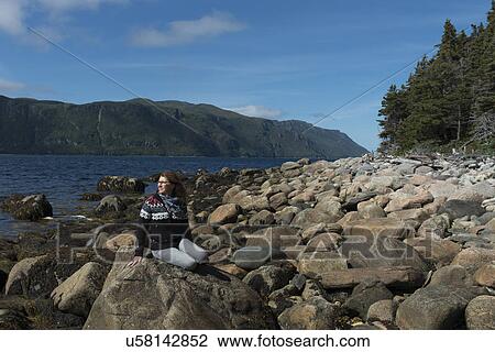 Stock Image - Woman sitting on rocks at coast, Norris Point, Bonne Bay, Gros Morne National Park, Newfoundland And Labrador, Canada. Fotosearch