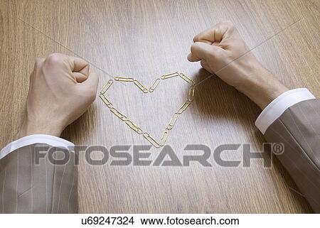 Picture - Business man clenching his fists with paper clip heart on desk. Fotosearch